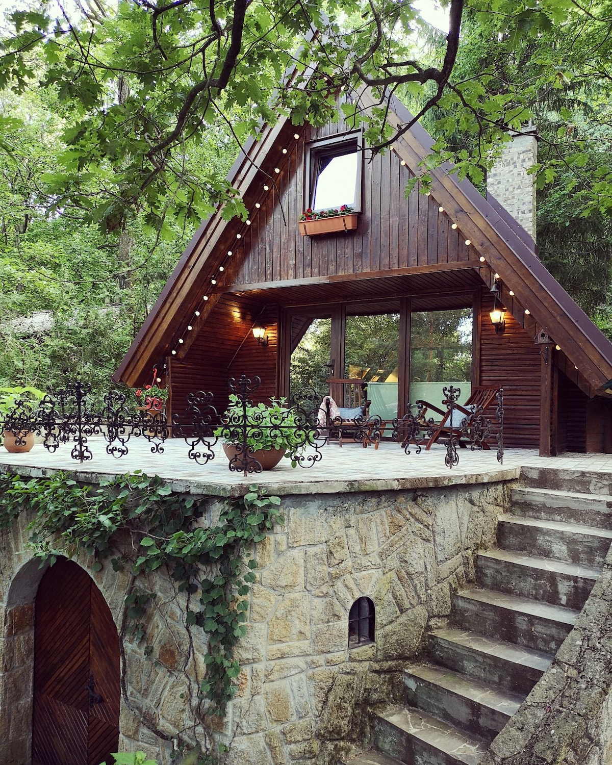 The charming cabin features a triangular roof and large glass windows, surrounded by lush greenery. A stone staircase leads to a spacious terrace adorned with decorative railings and potted plants, creating an inviting outdoor area for relaxation.