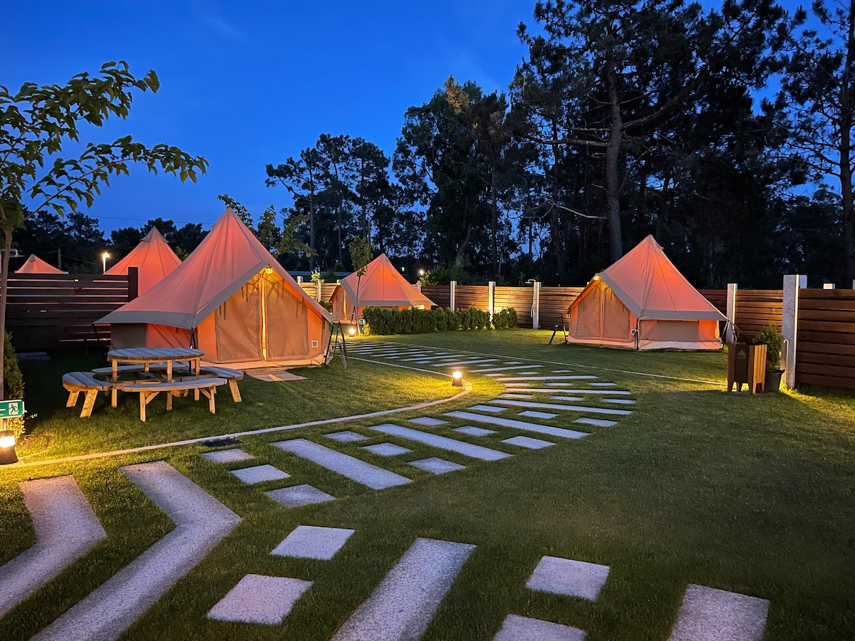 A serene glamping area is highlighted by tents arranged around a winding stone pathway. Soft lighting illuminates the space at twilight, surrounded by greenery and wooden fencing, creating an inviting outdoor atmosphere for relaxation.