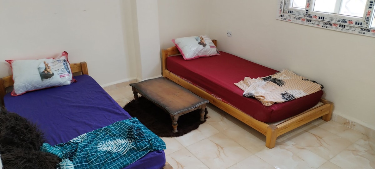 Two beds are positioned in a simple room, each with a colorful bedspread. A small wooden table occupies the center, accompanied by a plush rug underneath. Light enters through a window with decorative framing, enhancing the room's bright and open atmosphere.
