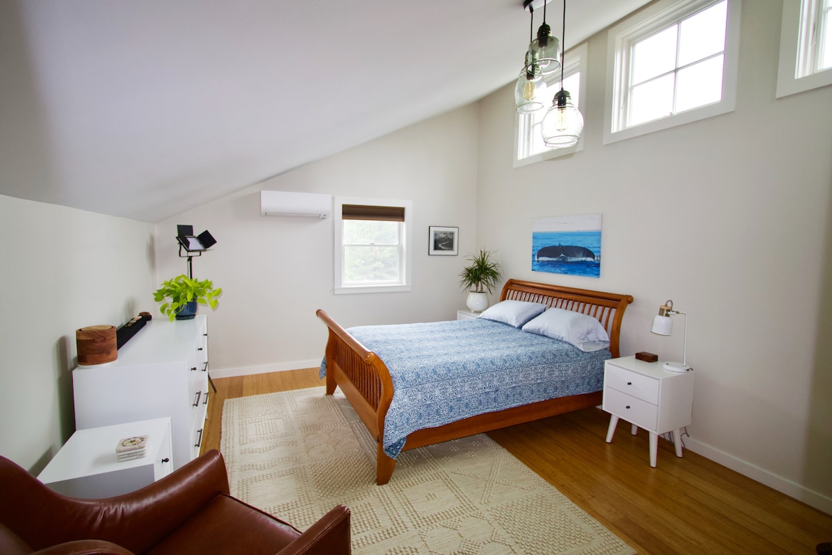 A cozy bedroom is highlighted by a wooden bed with a blue patterned bedspread. Natural light enters through three windows, illuminating the space. A potted plant and modern light fixtures enhance the atmosphere. A comfortable chair and minimalist storage add functionality.