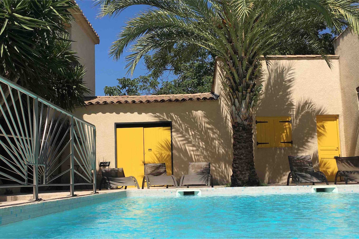 A swimming pool is set against a backdrop of lush greenery, with sun loungers positioned around the edge. Yellow doors can be seen on the adjoining structure, providing a vibrant contrast to the surroundings. Palm trees frame the scene, adding a tropical feel to the area.