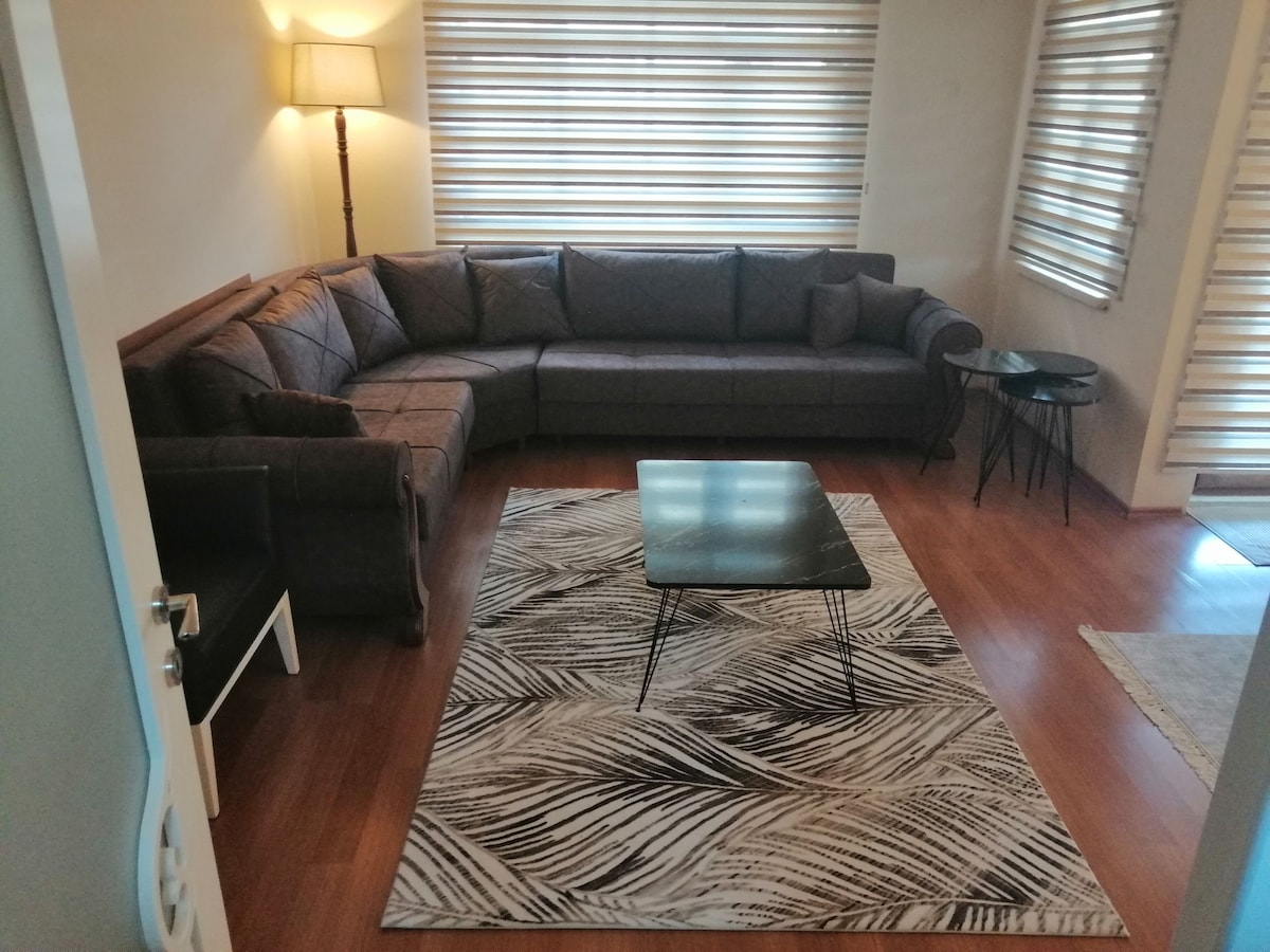 A spacious living area is furnished with a large sectional sofa arranged around a sleek coffee table, set atop a patterned area rug. Natural light filters through horizontal blinds, enhancing the warm tones of the wood flooring and creating a comfortable atmosphere.