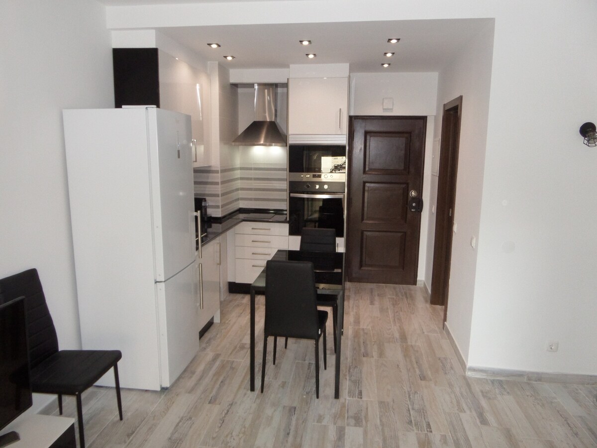 A modern kitchen is displayed, featuring sleek cabinetry and stainless steel appliances, including an oven and refrigerator. A dining area with four black chairs is situated nearby. The light-colored flooring complements the bright and airy space.