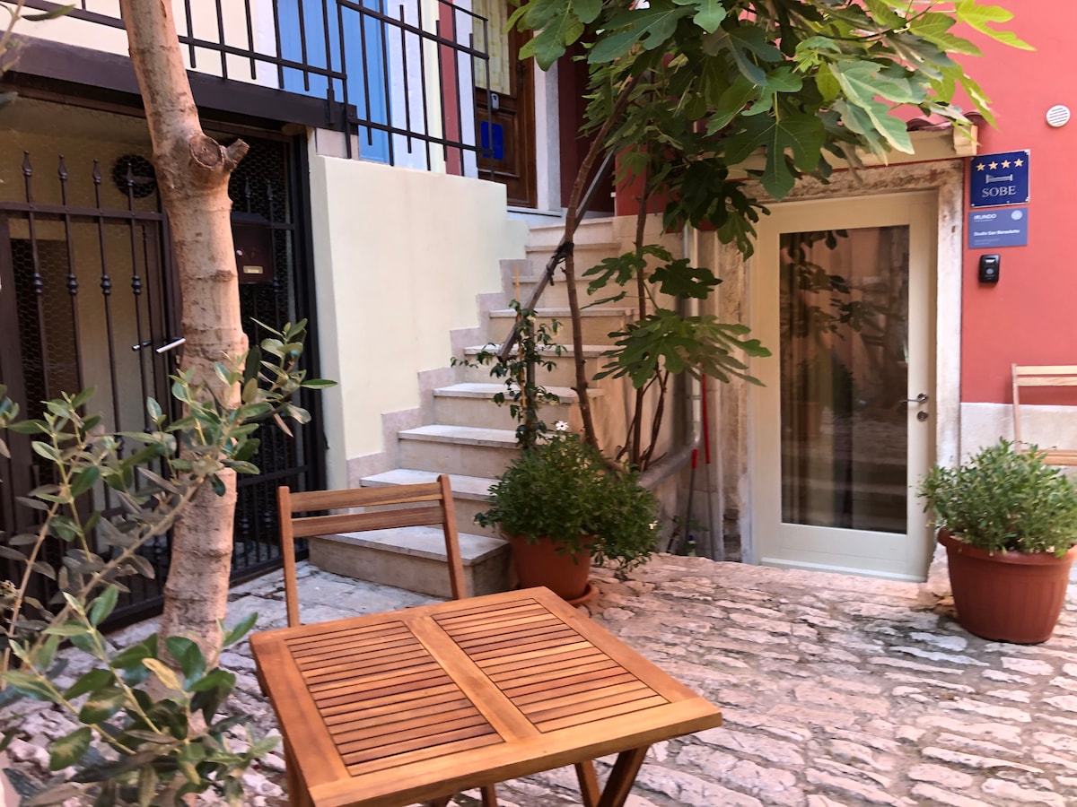 A stone-paved courtyard features a small wooden table paired with chairs, surrounded by potted plants. A staircase leads to an upper level, while a door is visible at the base of the stairs, framed by a vibrant wall.