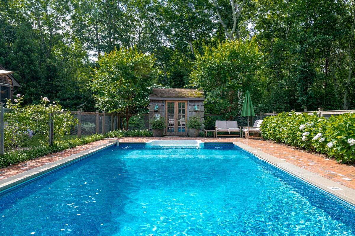 A heated pool is surrounded by lush greenery and features a rustic pool cottage in the background. Two chaise lounges are positioned beside the pool, offering a relaxing space under a large umbrella. Brick patio borders the water, creating a welcoming outdoor area.