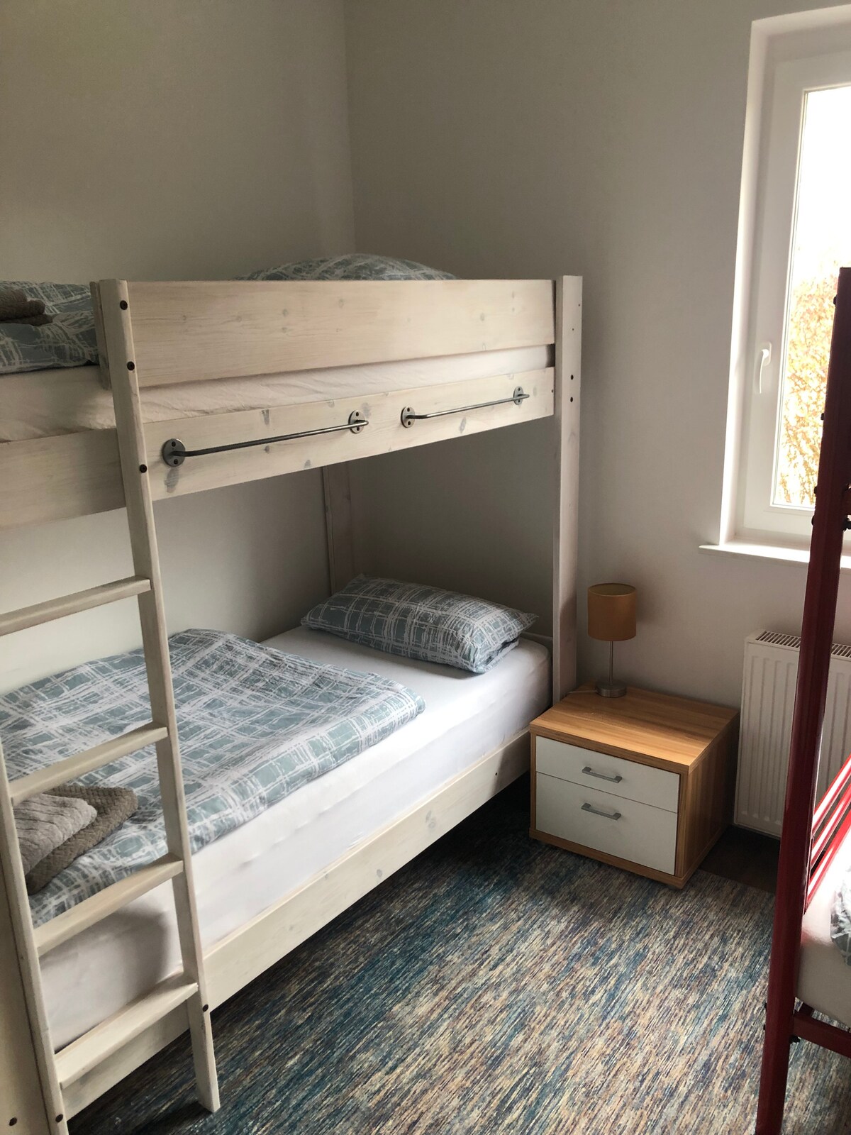 A cozy sleeping area features a wooden bunk bed with a light-colored finish. Soft bedding is arranged on both levels. A bedside table with a lamp stands next to the lower bunk. Natural light enters through a window, illuminating the space.