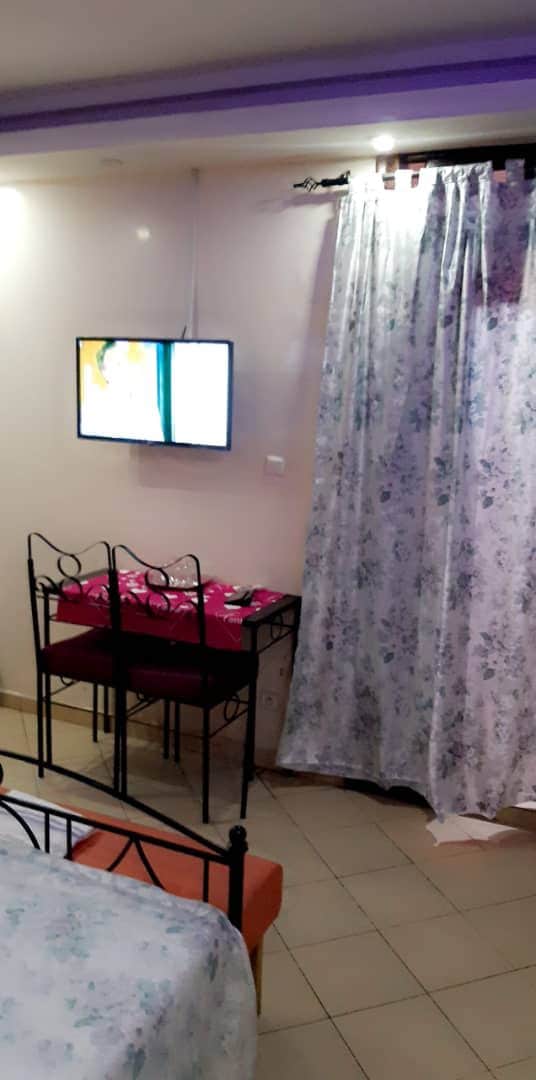 An independent room features a wall-mounted television and a small dining table with two chairs. A set of light-colored curtains adorned with floral patterns offers privacy. The tiled floor and warm wall colors contribute to a tidy, welcoming setting.