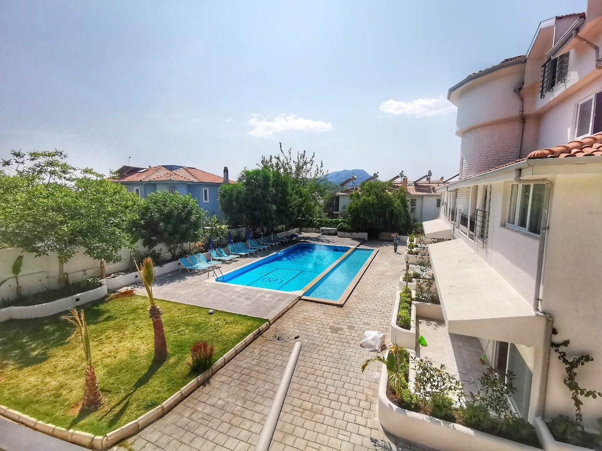 A sunny view of a private pool surrounded by lounge chairs and shaded areas. Lush greenery borders the pool area, enhancing the peaceful ambiance. The building features a spacious terrace, and nearby structures provide an unobstructed perspective of the pool and the garden.