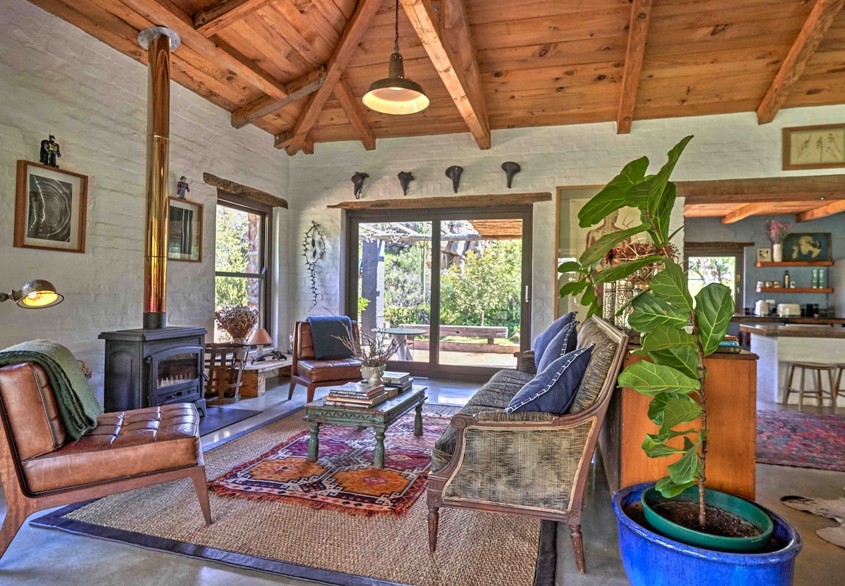 A warm living area features high wooden beams and a cozy fireplace. A rug lies beneath a coffee table surrounded by comfortable seating. Large sliding doors provide natural light and views of the outdoors, enhancing the connection with nature.