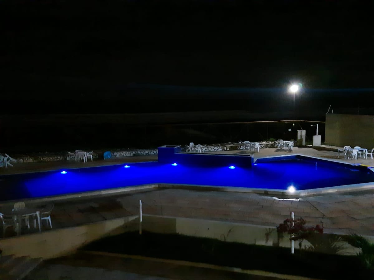 A tranquil pool area is illuminated by subtle blue lights against a dark backdrop. Multiple lounge chairs are arranged around the pool, and a building is visible in the background. The serene atmosphere suggests a peaceful retreat for relaxation.