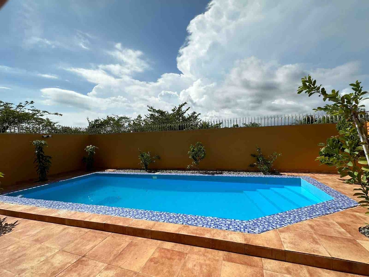A private pool is surrounded by a tiled patio, featuring gentle steps leading into the water. Lush greenery is visible along the edges, with a background of an expansive sky filled with clouds, creating a serene outdoor space.