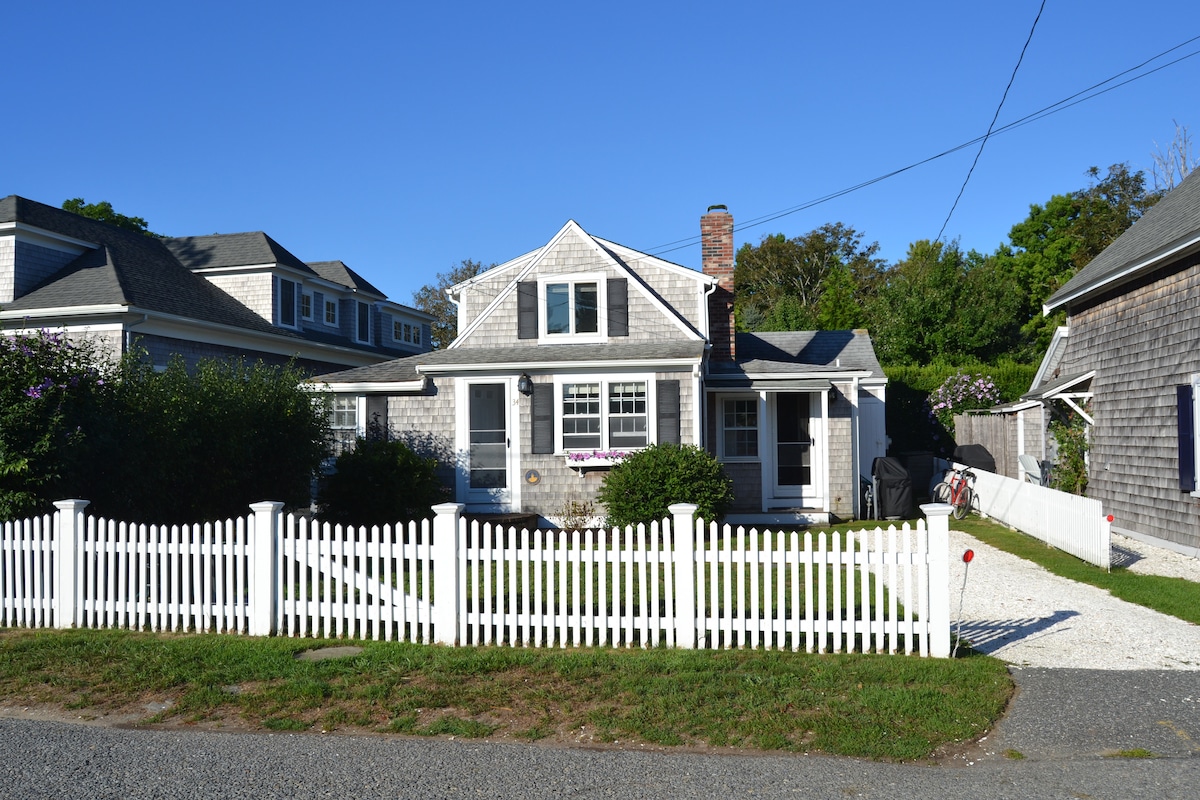 The exterior of the renovated Chatham home features a white picket fence and a gravel driveway. The house showcases a gabled roof and large windows, surrounded by greenery and trees. The clear blue sky above emphasizes the peaceful neighborhood setting.