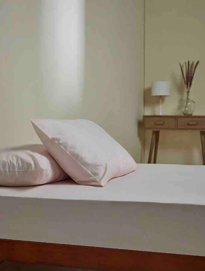 A simple sleeping area is presented with a spacious bed dressed in soft linens. Two pink pillows rest against the headboard. A small bedside lamp and decorative vase are visible on a wooden side table, creating an understated and calming space.