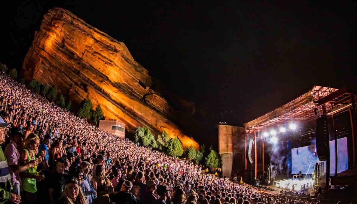 A lively concert scene is captured at Red Rocks, featuring a large audience gathered on stone steps. The stage, brightly lit, is framed by the towering red rock formations that provide a dramatic backdrop. Nighttime illuminates the setting, enhancing the atmosphere of the event.