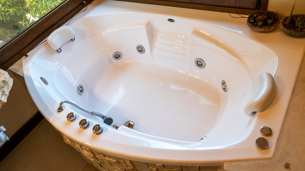 A spacious, inviting whirlpool bath is situated in the corner of the suite. The modern design features jets and smooth surfaces, with a view of greenery visible through the nearby window. Natural stone accents complement the bath area.