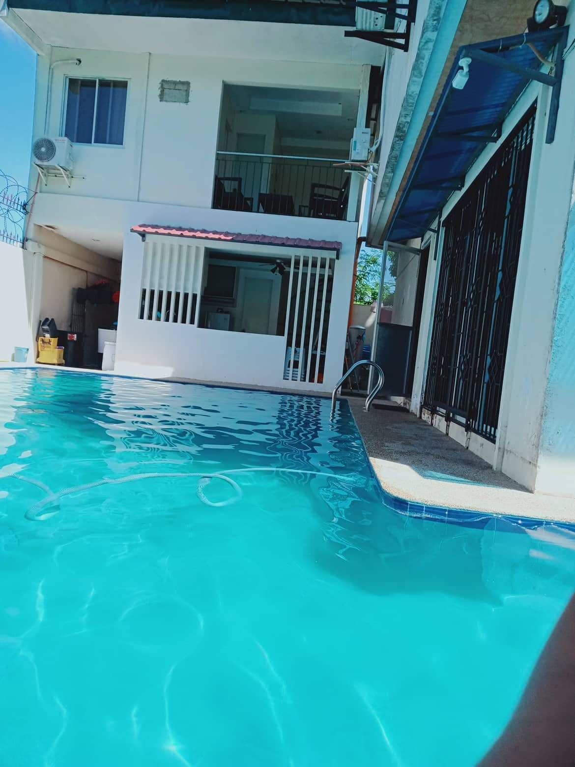 A clear blue swimming pool is viewed from the edge, reflecting sunlight. In the background, a two-story building is visible, featuring white walls and large windows. A staircase leads up to the second floor, while a small awning provides shade over an outdoor area.