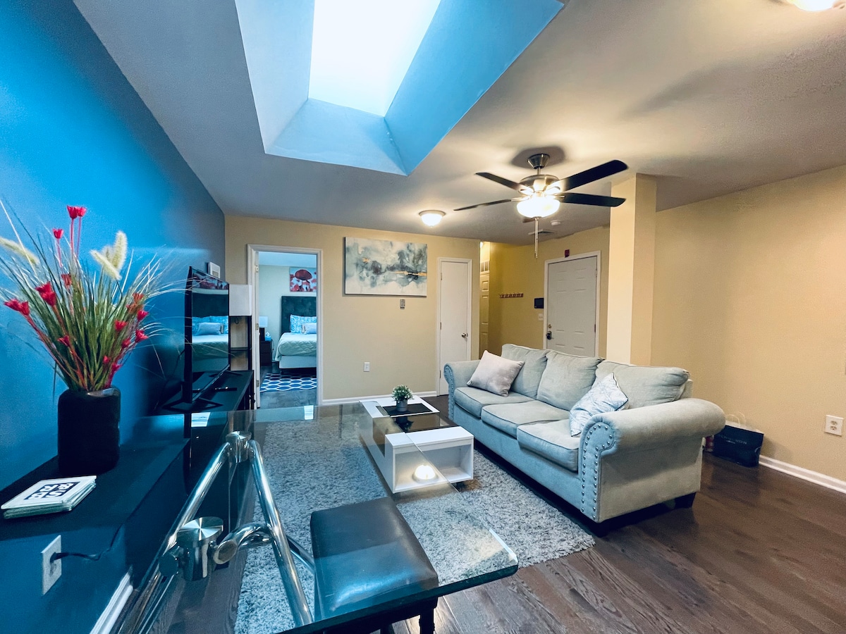 A spacious living area features a cozy light-colored sofa, a modern coffee table, and a sleek glass desk. A ceiling fan and skylight provide illumination, while a vibrant blue accent wall adds visual interest. Hardwood floors extend throughout the area, contributing to a welcoming ambience.