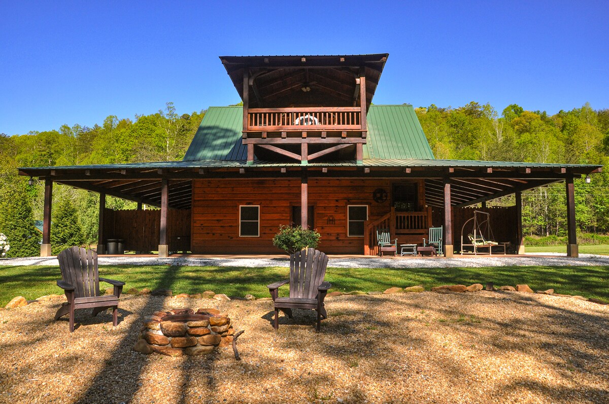A secluded cabin features a large, covered porch with multiple seating areas, ideal for relaxation. Wooden posts and a slanted green roof create an inviting exterior. A fire pit with stone seating provides a cozy gathering space, surrounded by lush greenery in the background.
