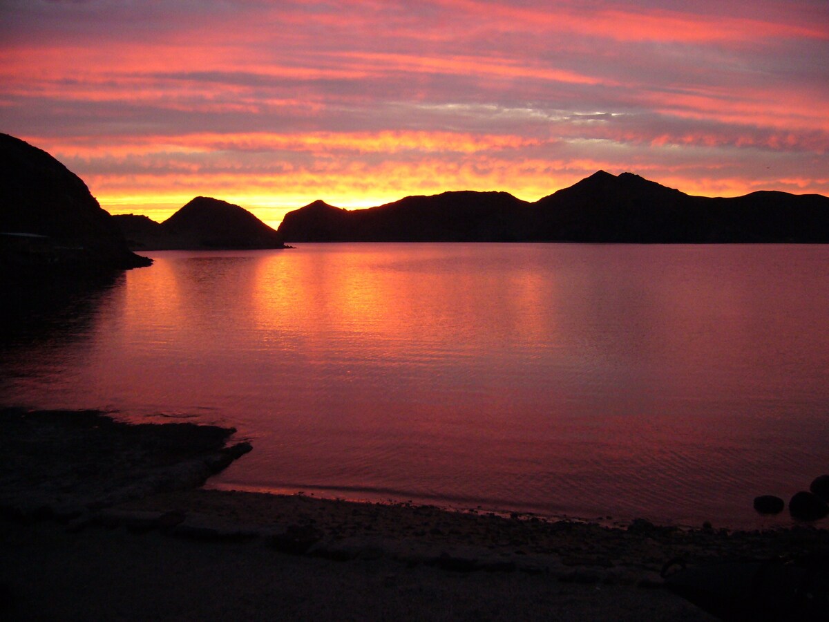 The image captures a serene sunset over the calm waters of the Sea of Cortez, with vibrant hues of orange, pink, and purple reflected on the surface. Silhouettes of mountains frame the horizon, adding to the tranquil scene. The shoreline is visible in the foreground.