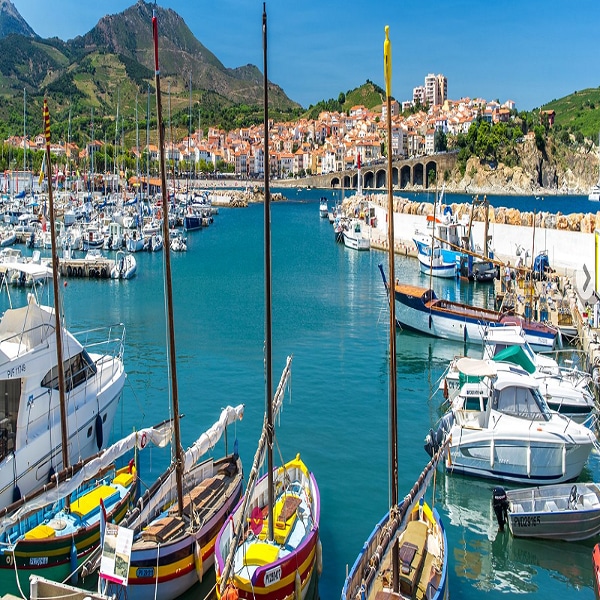 A vibrant harbor scene is portrayed, featuring colorful boats moored in tranquil waters. The backdrop reveals a picturesque village with buildings perched on a hillside, while lush green mountains rise in the distance under a clear blue sky.