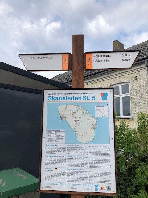 A directional signpost stands prominently, displaying distances to nearby locations, including Bäckviken and Norreborg. A map of the Skåneleden trail is attached, providing an overview of the hiking routes available in the area. The background features a modest building and green foliage.