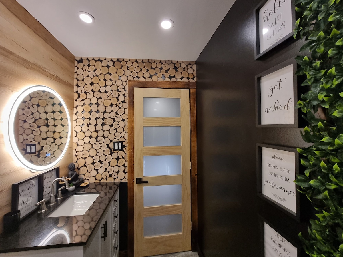 The bathroom features a modern design with a circular mirror illuminated by soft lighting. A wooden accent wall made of stacked logs contrasts with the sleek vanity and dark countertops. Framed art pieces are displayed on the walls, and green foliage adds a touch of nature.
