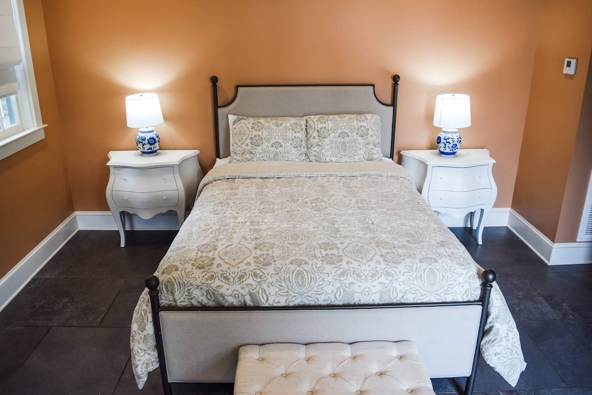 A cozy bedroom features a bed with soft bedding centered between two nightstands. Each nightstand is paired with a decorative lamp. The warm orange walls complement the neutral tones of the decor. Natural light enters through a window adorned with light blinds.