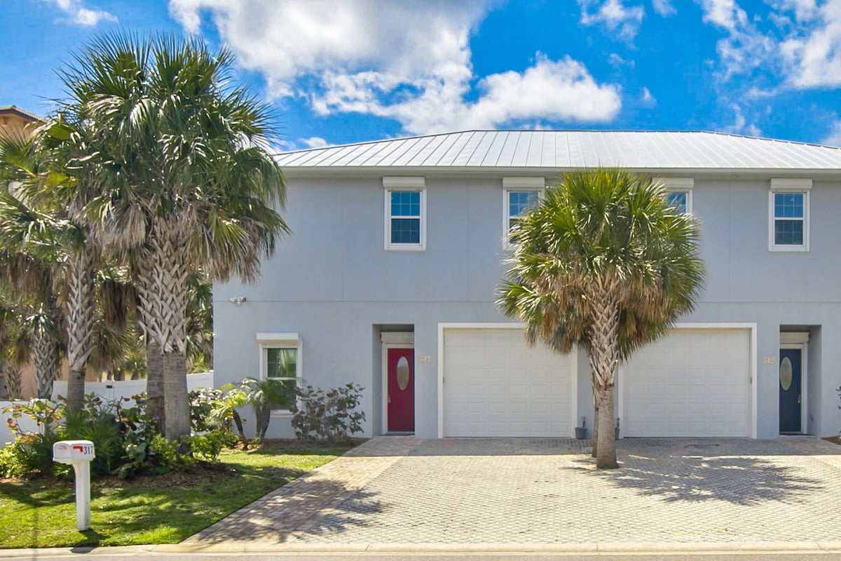 The exterior of a two-unit duplex is displayed, featuring a light gray facade and a modern metal roof. Palm trees and lush greenery frame the front, while two separate entrances are marked by round windows. A spacious driveway leads to two garages.
