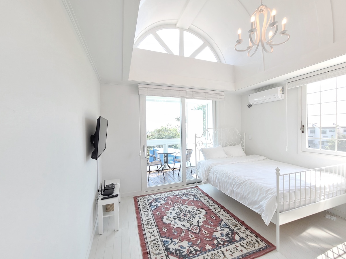 A bright and airy bedroom is highlighted by a spacious bed with white linens. Large glass doors open to a balcony, offering a view of the outdoors. A decorative chandelier hangs from the ceiling, and a flat-screen TV is mounted on the wall.