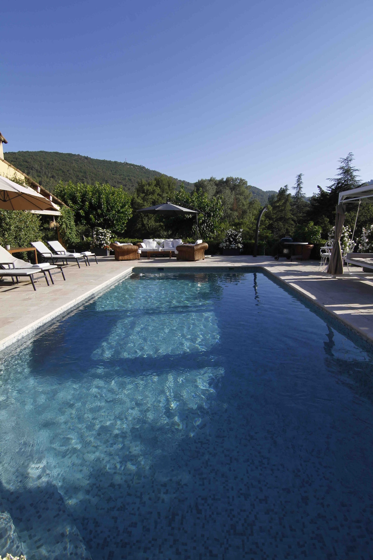 A serene swimming pool is framed by lounge chairs and shaded seating areas. Surrounding greenery includes trees and flowering plants, creating a peaceful outdoor setting. The clear water reflects the bright blue sky and distant hills, enhancing the tranquil ambiance of the villa's landscape.