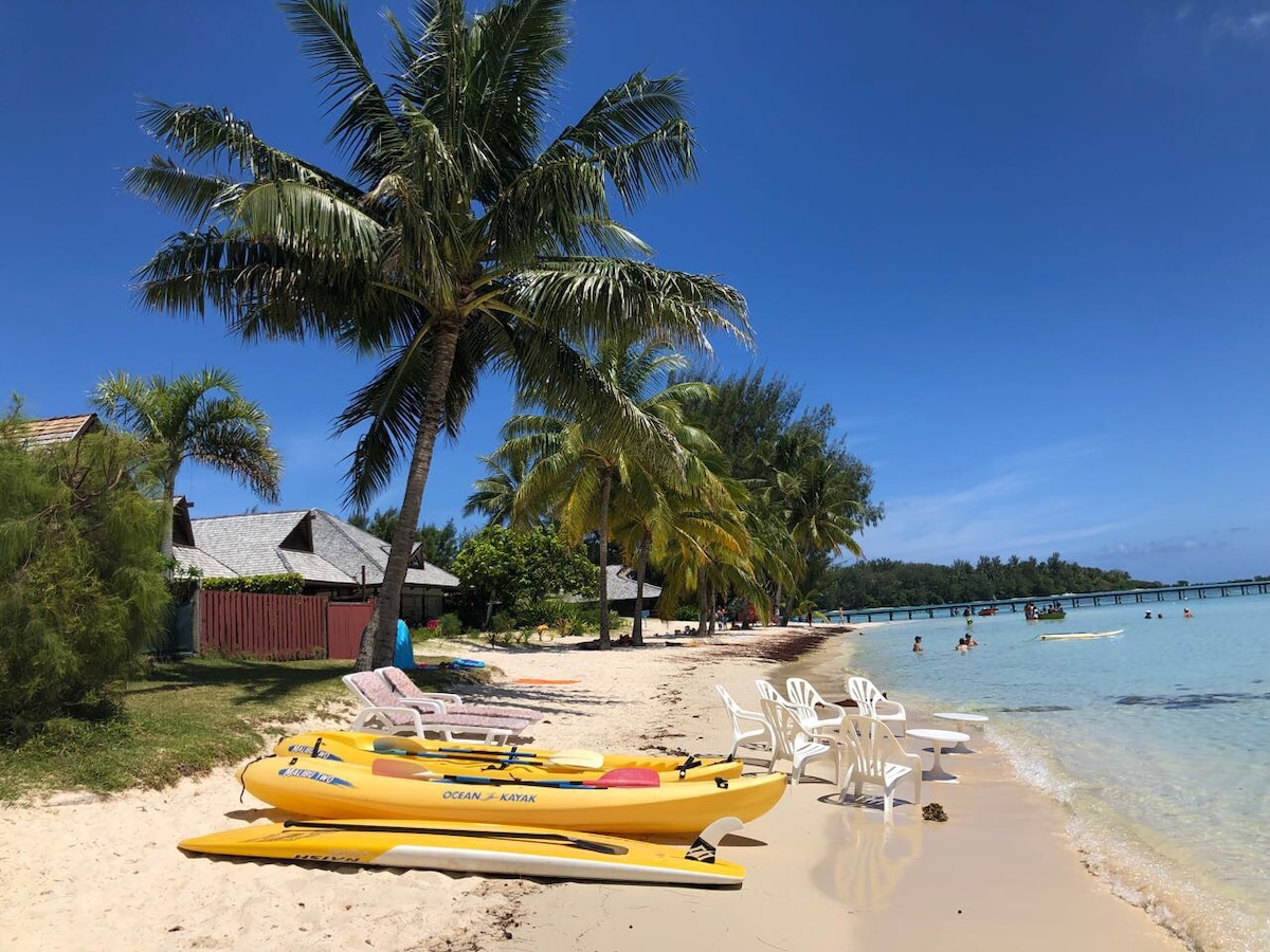 A sandy beach is lined with palm trees, providing shade beside the turquoise lagoon. Yellow kayaks rest on the shore, while several white chairs are arranged nearby. The clear sky reflects a serene atmosphere, with people enjoying the water further along the beach.