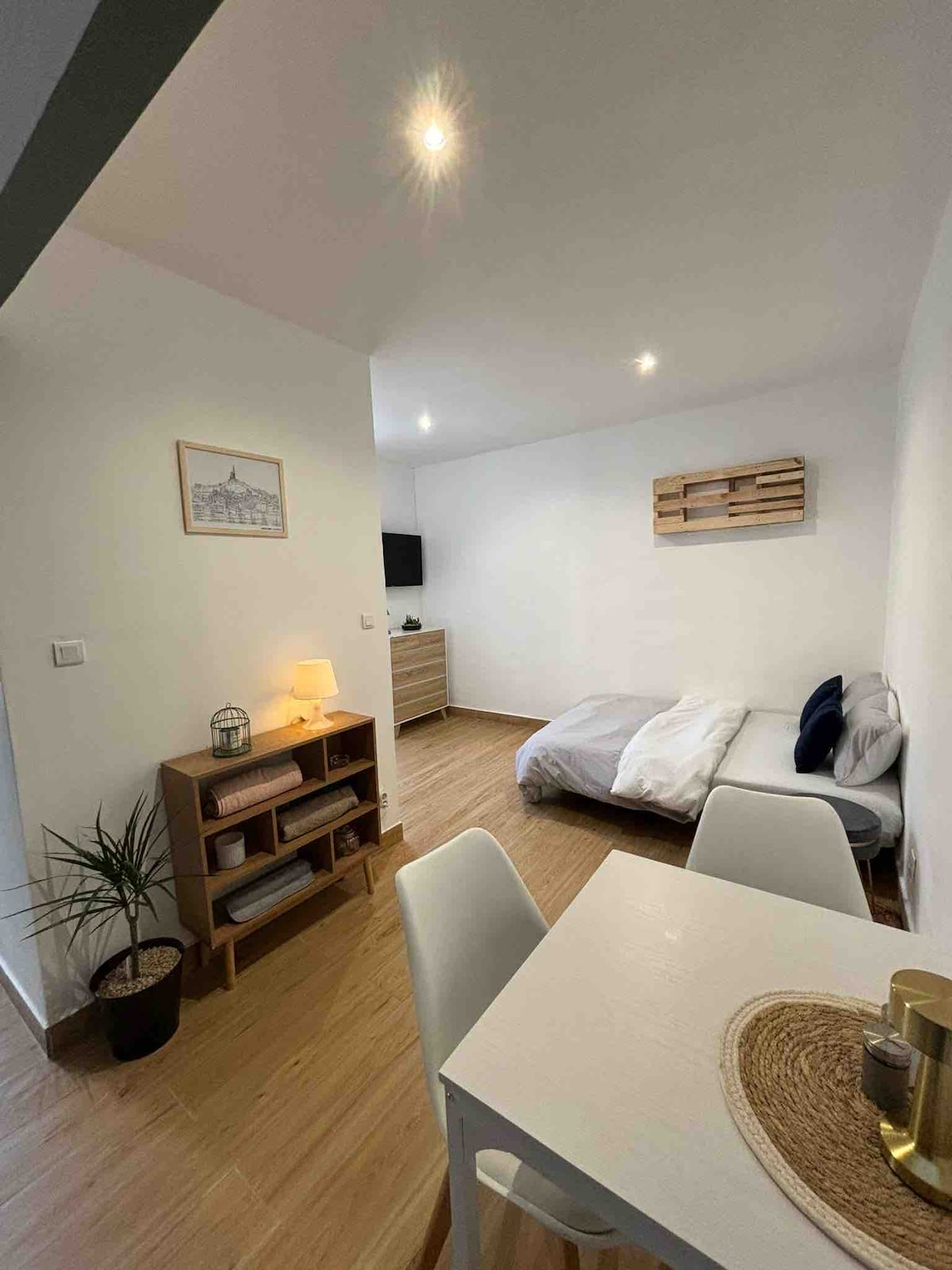 A well-lit studio space features a cozy sleeping area with a spacious bed, complemented by light wooden flooring. A small dining table with two chairs is present, alongside a shelf housing a decorative plant. A wall-mounted TV and neutral decor contribute to the minimalist ambiance.