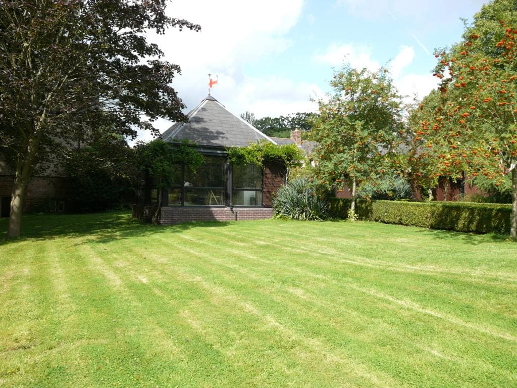 The exterior of the Garden Room is showcased, featuring a spacious lawn with neatly trimmed grass and a variety of trees. The building, with its distinctive roof, is framed by shrubs and plants, providing a serene setting. Bright natural light bathes the area.