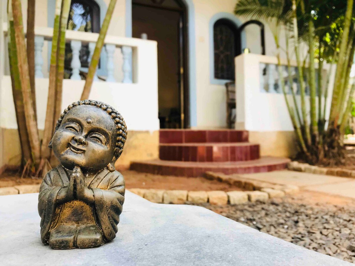 A serene Buddha statue is positioned on a stone surface in the foreground, with two palm trees flanking it and soft lighting enhancing its features. In the background, the entrance to a cozy home is visible, marked by soft arches and steps leading to the door.