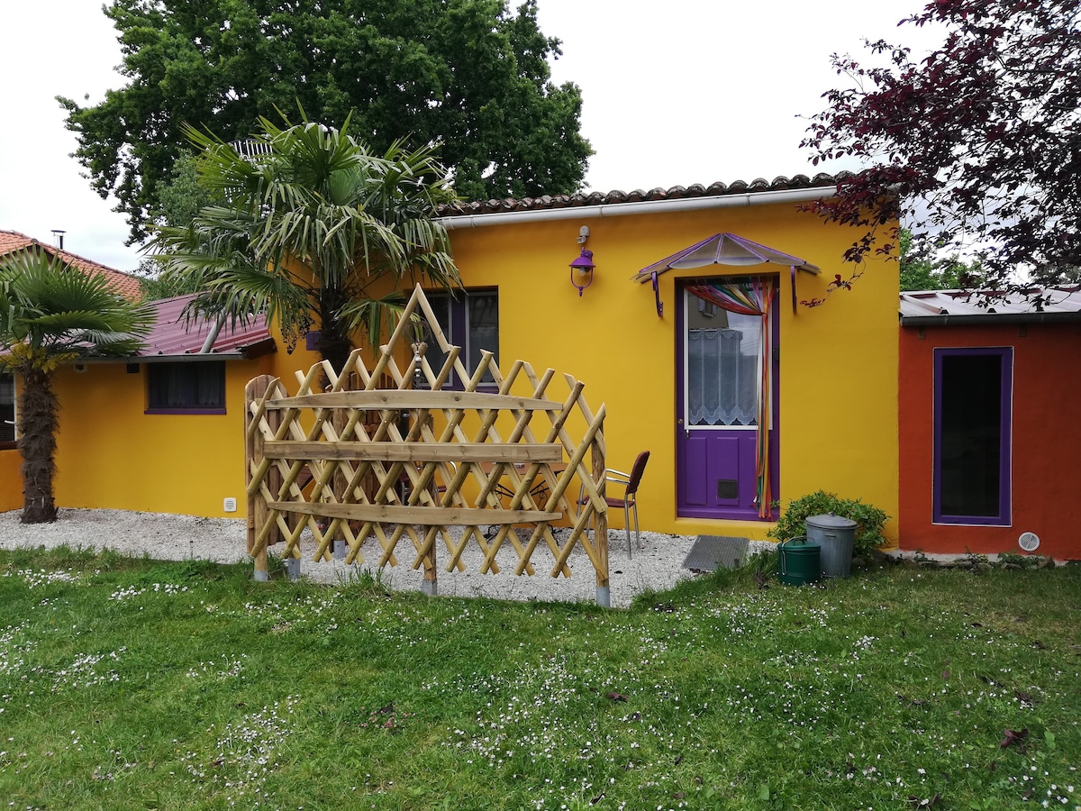 The image shows a charming yellow house adorned with purple accents and a welcoming front porch. A wooden fence surrounded by greenery offers privacy and a cozy outdoor space. Decorative elements include palm trees and colorful details near the entrance, enhancing the tranquil atmosphere.