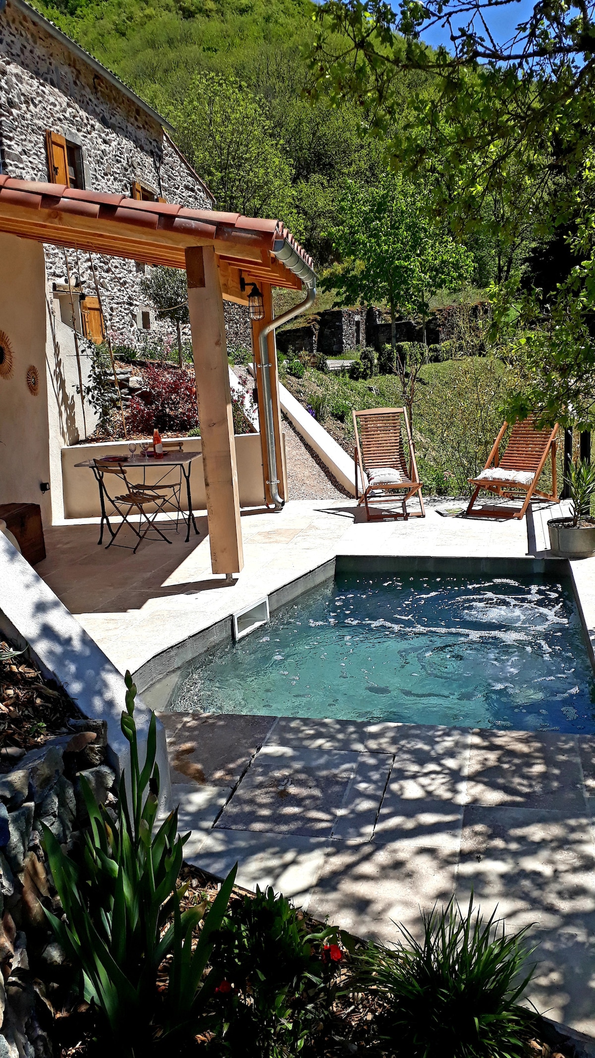 A private area features a small pool with bubbling water, surrounded by stone walls and lush greenery. Two lounge chairs are positioned under the shade of trees, while a compact dining table with chairs is nearby, ideal for enjoying outdoor meals.