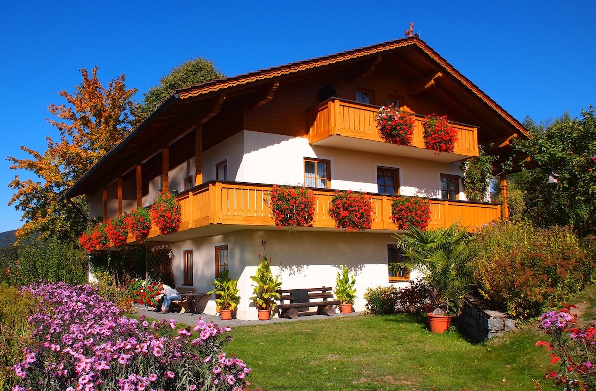 A two-story building is surrounded by vibrant gardens featuring flowers in various shades. The upper floors have wooden balconies with colorful flower boxes. A shaded outdoor seating area is located on the ground floor, providing a welcoming space amidst the greenery.