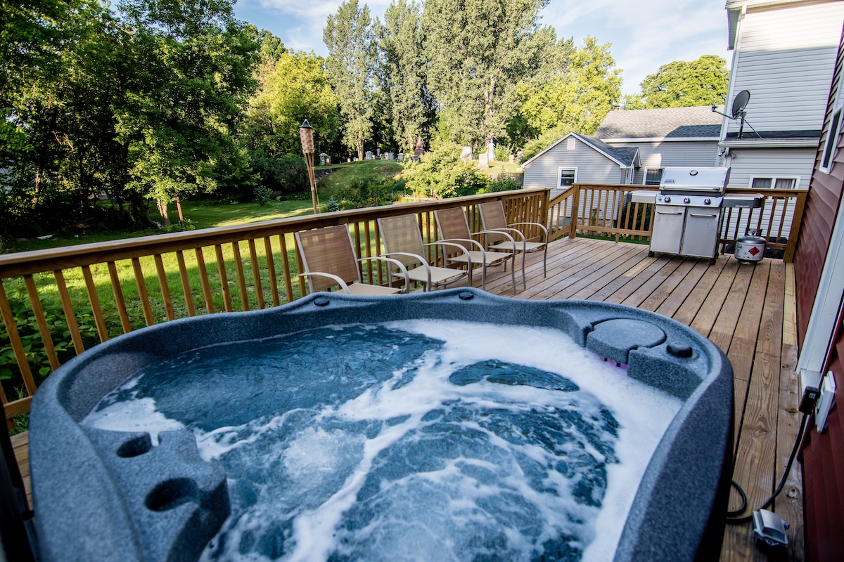A private back deck is presented, featuring a four-person Jacuzzi with bubbling water. Surrounding seating options, including several chairs and a grill, provide ample space for outdoor gatherings, all overlooking a grassy lawn and wooded area in the background.