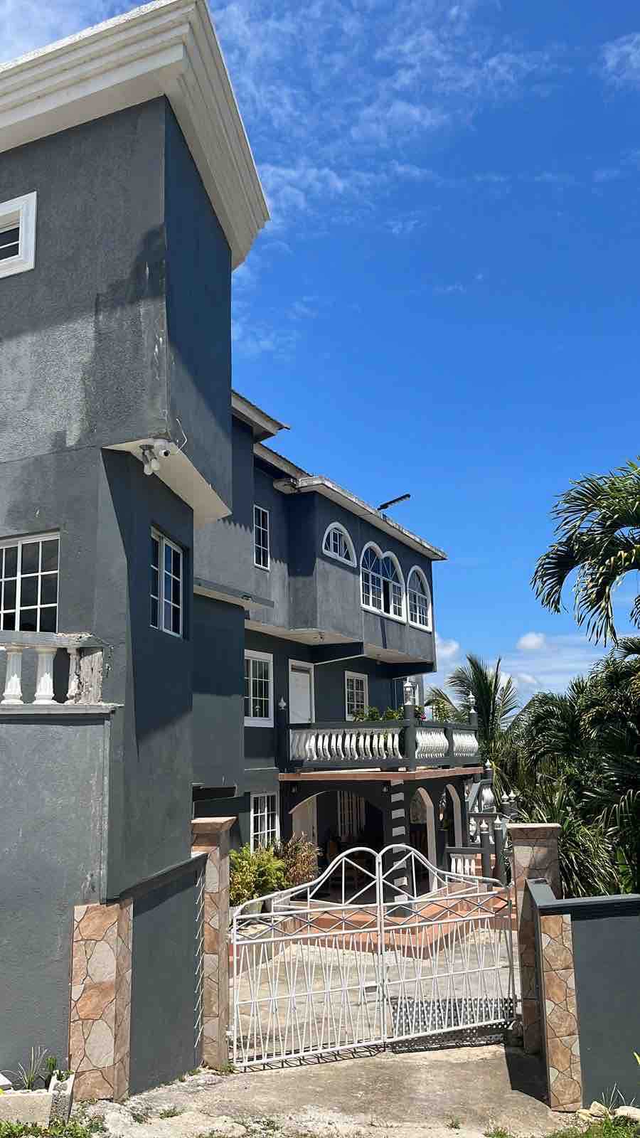 The exterior of a multi-story building is showcased, featuring large arched windows and a decorative balcony. A gated entrance leads to a landscaped area with palm trees. The bright blue sky creates a contrasting backdrop, highlighting the building's architecture and surroundings.