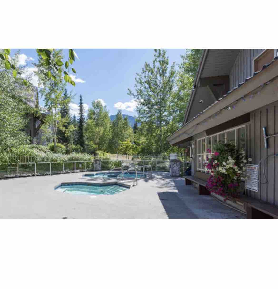 An outdoor area features multiple hot tubs surrounded by lush greenery and colorful floral arrangements. The building's exterior includes large windows that provide natural light. A paved space is visible, offering a tranquil setting for relaxation.