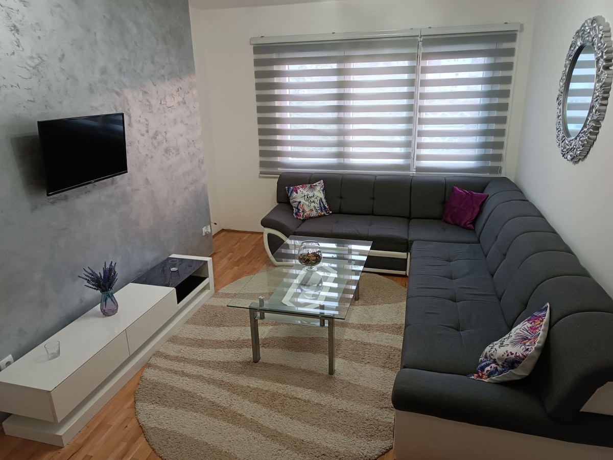 A modern living area is equipped with a sectional sofa and a glass coffee table, positioned on a round rug. Natural light filters through large windows with stylish blinds, and a wall-mounted television is visible. Decorative pillows add subtle color to the seating arrangement.