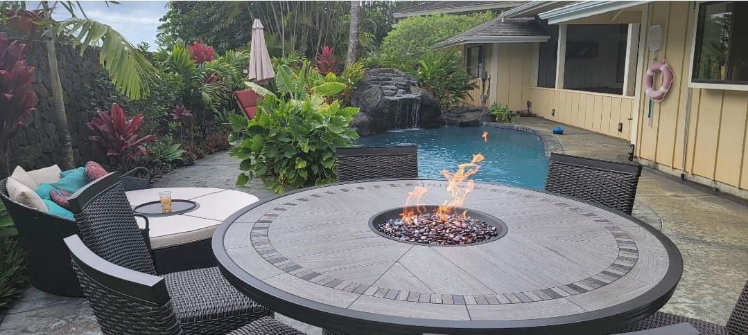 An outdoor seating area features a round table with a fire pit at its center, surrounded by comfortable chairs. Lush tropical foliage frames a private salt water pool with a waterfall, creating a serene setting for relaxation.