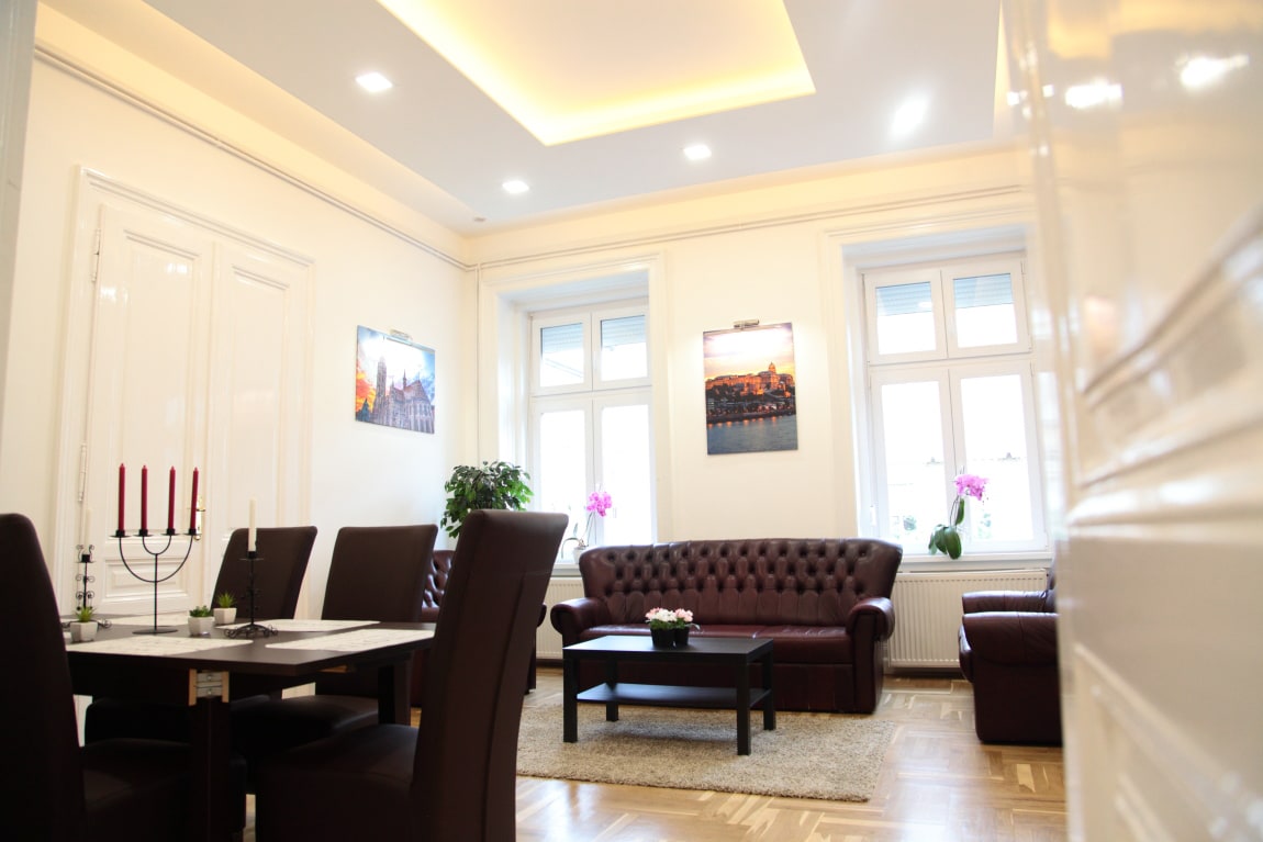 An elegantly furnished living area features a brown leather sofa and matching armchairs arranged around a dark wood coffee table. Light streams in through large windows, enhancing the room's brightness, while framed photographs adorn the walls. A dining table for four is positioned nearby.