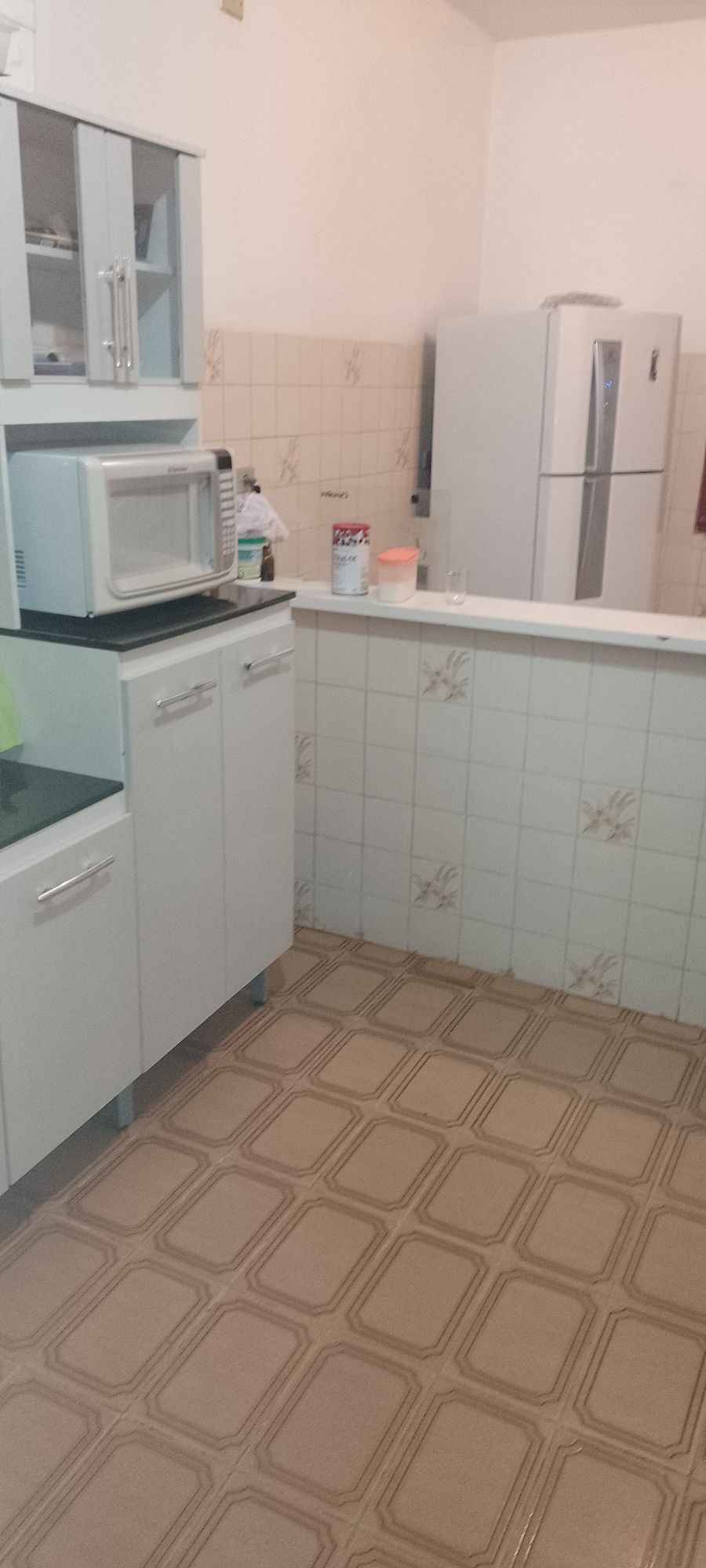 A functional kitchen space is shown, featuring white cabinets and a countertop. A microwave stands on the counter, along with various kitchen supplies. A refrigerator can be seen in the background, and the tiled floor adds a classic touch to the area.
