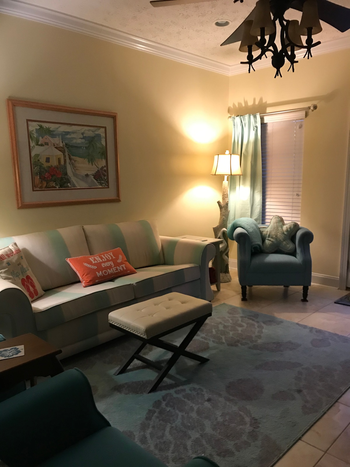 A cozy living space is presented with soft pastel colors, featuring a striped sofa and a comfortable armchair. A decorative painting is hung on the wall, and warm light is provided by a floor lamp. A neutral-toned rug adds texture to the tiled floor.
