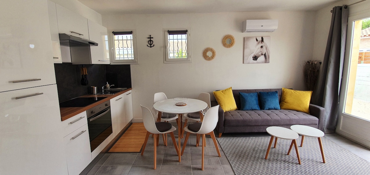 A modern living area features a gray sofa accompanied by two small tables. A round dining table with four chairs is positioned nearby. The kitchen area is equipped with sleek cabinetry and appliances. Natural light enters through the windows, creating a bright atmosphere.