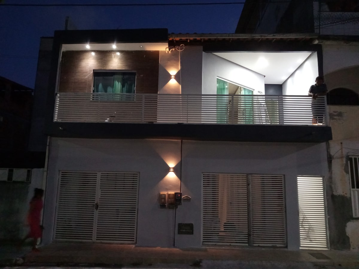 The exterior of a two-story apartment building is shown at dusk. Modern architectural features include a balcony with glass railing and warm white exterior lighting. Large glass doors are displayed at the ground level, enhancing visibility into the interior space.