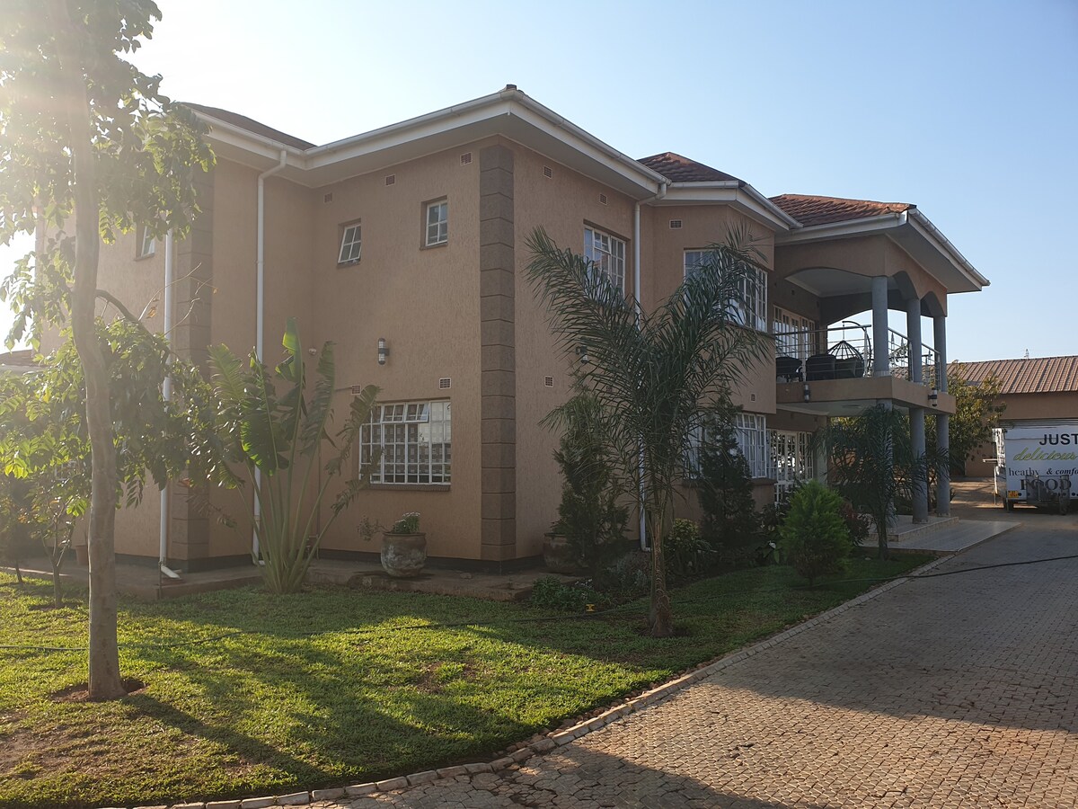 A spacious two-story house is set amidst green lawns and palm trees. The exterior features large windows that invite natural light, while a covered veranda overlooks the yard. The structure is framed by a paved driveway, ensuring easy access.