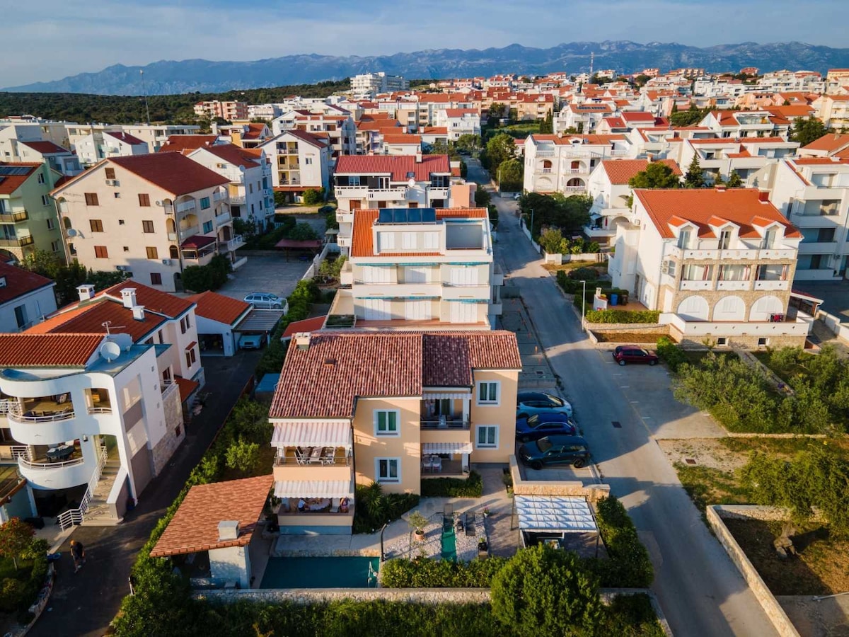 An aerial view showcases a residential area with a mix of low-rise buildings and villas, characterized by terracotta roofs. The apartment building is highlighted, surrounded by greenery and a few parked cars. The backdrop features distant mountains against a clear sky.