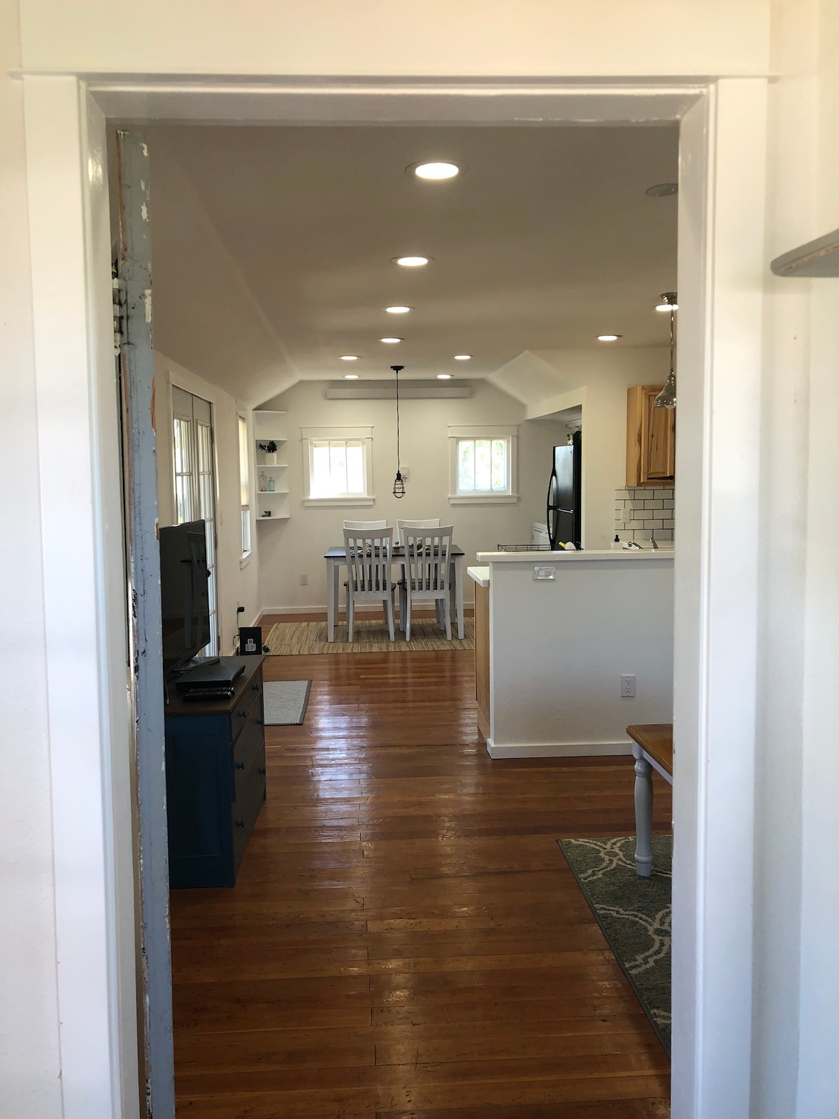 A hallway extends towards a light-filled kitchen area containing a dining table with four chairs. Warm wooden floors contrast with white cabinetry and walls, while soft lighting above enhances the open layout. A cozy area rug lies at the base of the table, adding texture to the space.